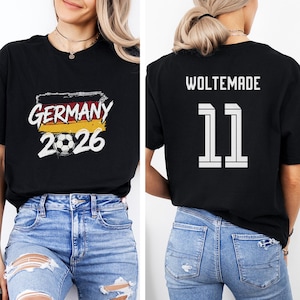Puede incluir: Camiseta negra con el texto "GERMANY 2026", un gráfico de balón de fútbol y los colores de la bandera alemana. La parte trasera muestra "WOLTEMADE" y el número "11" en blanco.