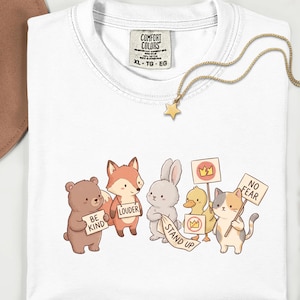 Könnte beinhalten: Weißes Comfort Colors T-Shirt mit einer Grafik von Zeichentricktieren, die Schilder mit den Aufschriften "Be Kind", "Louder", "Stand Up" und "No Fear" halten. Eine goldene Stern-Anhänger-Halskette liegt auf dem T-Shirt.
