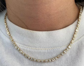 May include: A gold chain necklace featuring a repeating pattern of bezel-set clear stones in various shapes, including round, marquise, pear, and emerald cuts. The necklace rests against a white fabric surface, showcasing a continuous metallic link design.