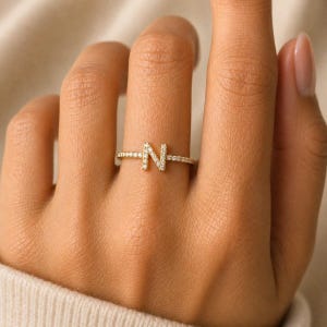 Puede incluir: Un anillo fino de oro que presenta una letra N mayúscula como pieza central. Tanto la banda como la letra están incrustadas con pequeñas piedras claras y brillantes que cubren toda la superficie del anillo, creando un efecto de brillo continuo.