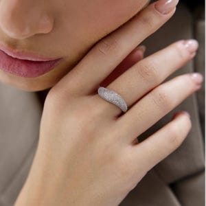 Könnte beinhalten: Ein silberfarbener Ring mit einer breiten, sich verjüngenden Ringschiene, die vollständig mit kleinen, funkelnden klaren Steinen besetzt ist. Der Ring hat eine glatte, abgerundete Oberfläche, die sich leicht wölbt und eine Kuppelform bildet.