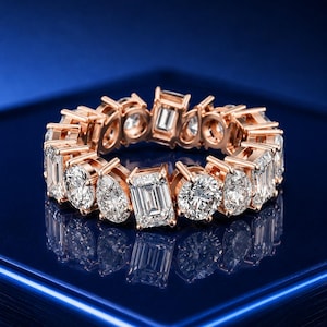 May include: A rose gold eternity band featuring an alternating pattern of clear, faceted emerald-cut and round-cut stones set in a continuous circle. The metal has a warm, metallic finish, and the stones are arranged to create a repeating sequence.
