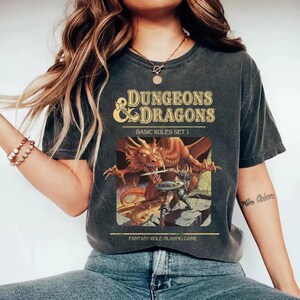Op de afbeelding: Een antracietgrijs t-shirt met korte mouwen en een Dungeons & Dragons Basic Rules Set 1-afbeelding. De print toont een grote oranje draak tegenover een krijger met een zwaard en schild, met de tekst Dungeons & Dragons en Fantasy Role-Playing Game.