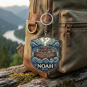 May include: A clear acrylic keychain with a colourful illustration of Noah's Ark. The design includes the ark, animals, clouds, and the word "NOAH" with "EST. B.C." below. The keychain is attached to a silver chain and ring.