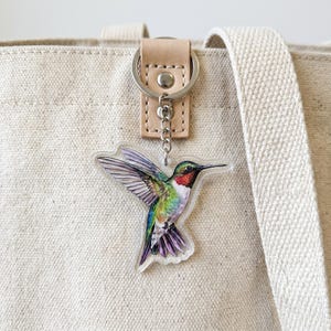 May include: A vibrant hummingbird keyring with a silver ring and a tan leather strap. The hummingbird features green, blue, red, and purple feathers. The keyring is attached to a beige canvas tote bag.