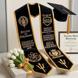 May include: Black graduation sash with gold trim and text, "Doctor of Philosophy" and "Class of 2024." Includes a mortarboard cap, diploma, and a bouquet of white tulips. The sash features the Arizona State University logo.