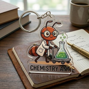 May include: A clear acrylic keychain featuring a cartoon ant in a lab coat pushing a cart with a green liquid-filled flask. The keychain has the words "CHEMISTRY ANT" printed on it. The keychain is attached to a silver keyring.