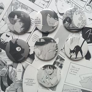 Könnte beinhalten: Sieben runde Ansteckbuttons mit schwarz-weißen Illustrationen im Manga-Stil. Die Motive zeigen Charaktere mit ausdrucksstarken Gesichtern, eine Katzensilhouette und eine Katze mit gekrümmtem Rücken. Einige Buttons enthalten französische Sprechblasen.