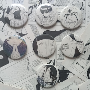 May include: Seven circular pin-back buttons featuring black and white manga character illustrations. One button displays a holographic finish with a character in a prayer pose. Another button includes the French text COMME DE VRAIS SHINOBIS.