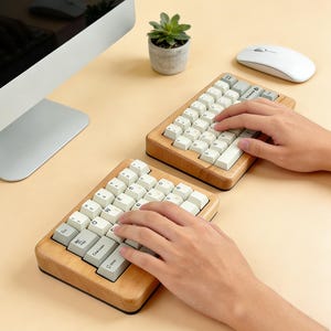 Könnte beinhalten: Zwei ergonomische Holz-Tastaturen mit cremefarbenen Tastenkappen sind auf einer beigefarbenen Oberfläche zu sehen. Ein Computermonitor, eine weiße Maus und eine kleine Topfpflanze sind ebenfalls sichtbar. Die Tastaturen werden benutzt.