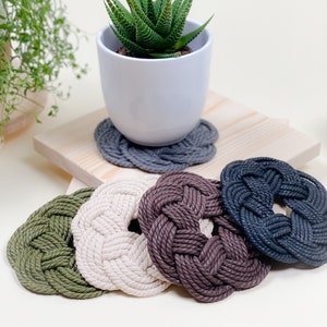Könnte beinhalten: Fünf runde Seiluntersetzer in verschiedenen Farben: Grün, Beige, Braun, Grau und Schwarz. Die Untersetzer sind aus dickem Seil gefertigt und haben ein Knotendesign. Eine kleine Topfpflanze sitzt auf dem grauen Untersetzer.