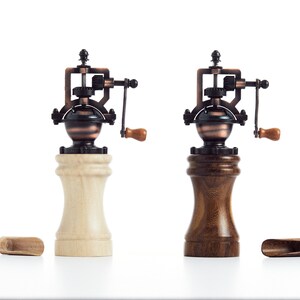 May include: Two vintage-style manual pepper grinders with wooden bases. One grinder has a light wood base and the other has a dark wood base. Both grinders have a copper and black metal mechanism with a crank handle.