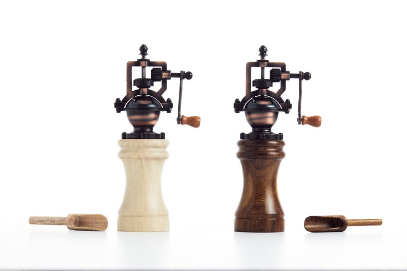 May include: Two vintage-style manual pepper grinders with wooden bases. One grinder has a light wood base and the other has a dark wood base. Both grinders have a copper and black metal mechanism with a crank handle.