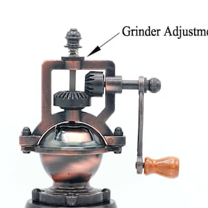 May include: A vintage-style manual coffee grinder with a copper finish. The grinder has a crank handle and a gear system for grinding coffee beans. The grinder has a knob on top for adjusting the grind size. The text "Grinder Adjustment" is visible in the image.
