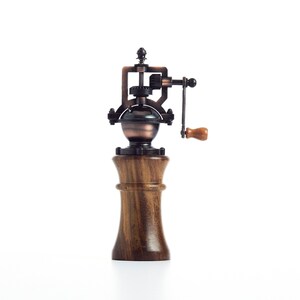 May include: A vintage-style wooden pepper mill with a metal grinder and a hand crank. The mill is made of dark wood and has a copper-colored metal top.
