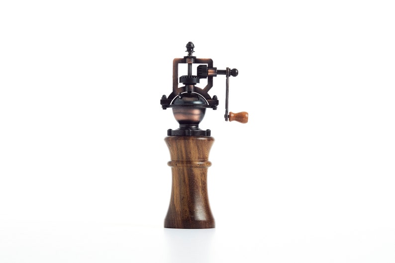 May include: A vintage-style wooden pepper mill with a metal grinder and a hand crank. The mill is made of dark wood and has a copper-colored metal top.