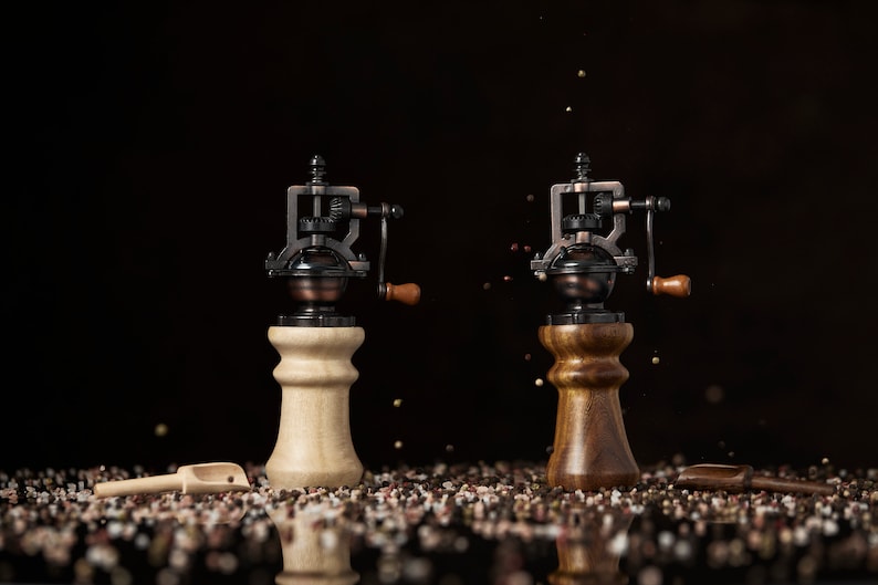 May include: Two antique-style pepper grinders with wooden bases. One grinder has a light wood base and the other has a dark wood base. Both grinders have a black metal mechanism and a small wooden handle.