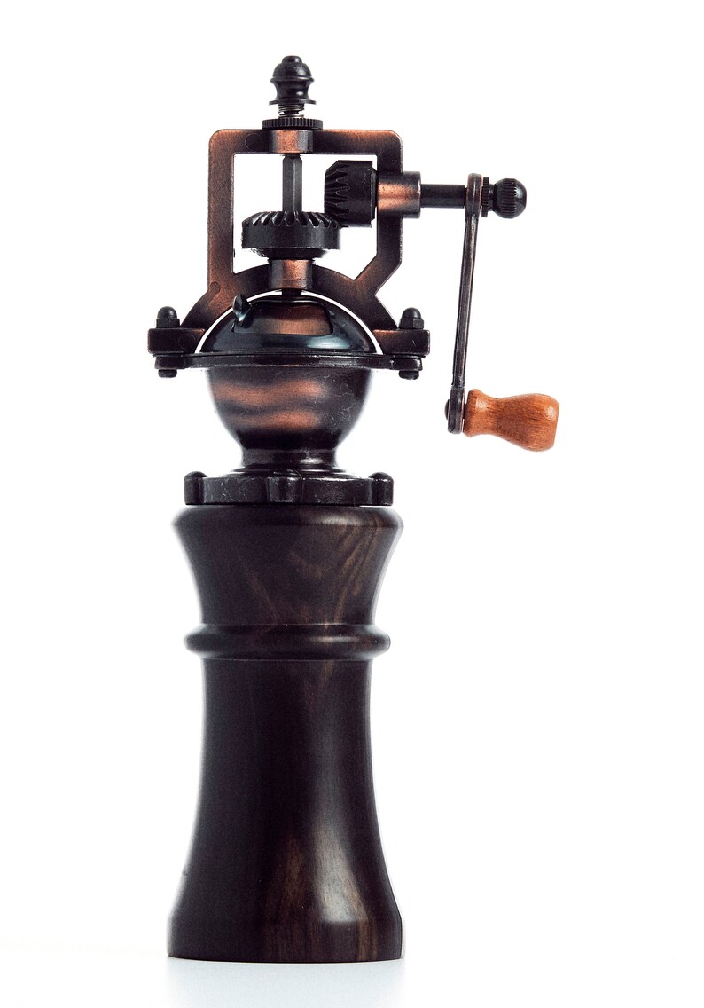 May include: A vintage-style manual coffee grinder with a wooden base and a copper-toned metal top. The grinder has a crank handle and a gear system visible on the top.
