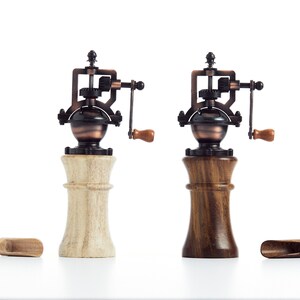 May include: Two antique-style salt and pepper grinders with wooden bases. One base is light wood and the other is dark wood. Both grinders have a copper-colored metal top with a crank.