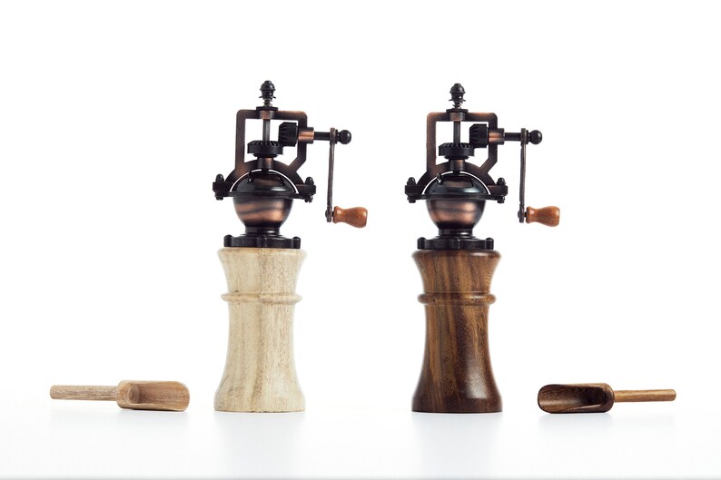 May include: Two antique-style salt and pepper grinders with wooden bases. One base is light wood and the other is dark wood. Both grinders have a copper-colored metal top with a crank.