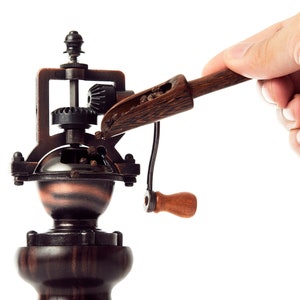 May include: A vintage-style wooden pepper grinder with a hand crank and a scoop filled with peppercorns. The grinder is made of dark wood and metal with a copper-colored base.