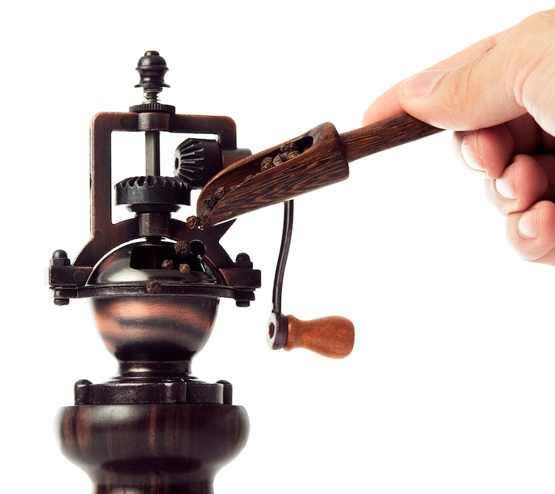 May include: A vintage-style wooden pepper grinder with a hand crank and a scoop filled with peppercorns. The grinder is made of dark wood and metal with a copper-colored base.