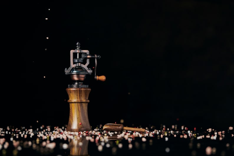 May include: A vintage wooden pepper grinder with a metal mechanism and a crank handle. The grinder is surrounded by a scattering of black, white, and pink peppercorns on a black surface.