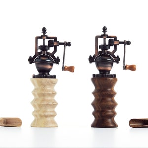 May include: Two antique-style manual coffee grinders with wooden bases. One base is light wood and the other is dark wood. Both grinders have a copper and black finish.