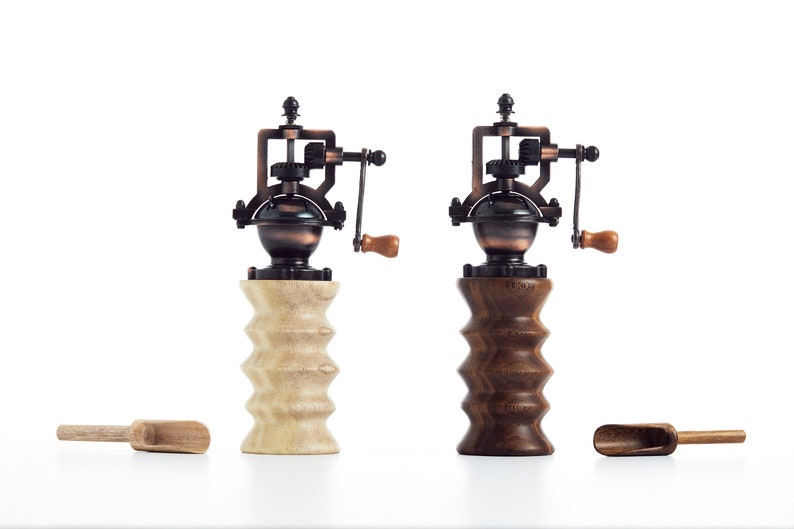 May include: Two antique-style manual coffee grinders with wooden bases. One base is light wood and the other is dark wood. Both grinders have a copper and black finish.
