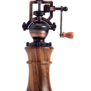 May include: A vintage-style wooden pepper mill with a metal grinder and a hand crank. The grinder is made of copper and has a gear system. The mill is made of dark wood with a grain pattern.