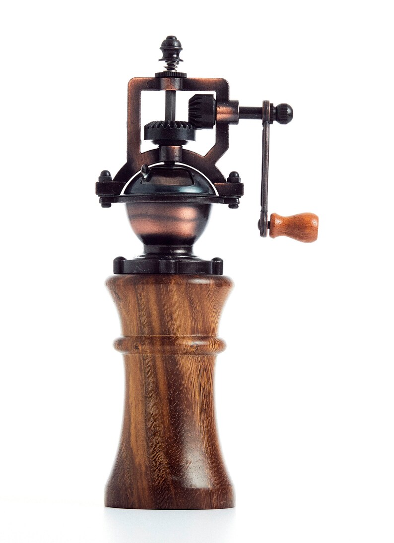 May include: A vintage-style wooden pepper mill with a metal grinder and a hand crank. The grinder is made of copper and has a gear system. The mill is made of dark wood with a grain pattern.