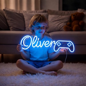 May include: A blue neon sign in the shape of a video game controller with the name "Oliver" in cursive script. The sign is held by a child sitting on a white rug. The sign is illuminated with a bright blue light.