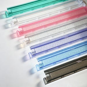 May include: A selection of colourful, transparent plastic organisers in shades of green, white, pink, silver, blue, and black. Each organiser has a rectangular shape with a small handle on one end. The organisers are arranged diagonally against a white background.