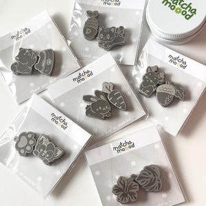May include: Six sets of grey enamel pins on branded matcha mood backing cards. Designs include a cat with a fish bone, a rabbit with a carrot, a paw print with a cat face, a deer with an acorn, a flower with a leaf, and a dog with a food bowl.
