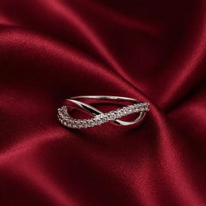 Puede incluir: Un anillo plateado con forma de infinito, adornado con pequeñas piedras preciosas transparentes. Se encuentra sobre un fondo de satén rojo intenso, creando una estética lujosa y elegante. El diseño del anillo presenta un bucle continuo, que simboliza la eternidad.