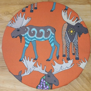 May include: Circular orange fabric featuring a repeating pattern of stylised moose. The moose have large antlers and are decorated with various designs, including waves, stripes, and floral patterns. The moose are brown, blue, and white.