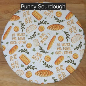 May include: A circular bread cover made of white fabric, adorned with illustrations of various breads in brown and yellow. The design includes text such as "Punny Sourdough", "You can crust me", "I loaf you", and "At yeast we have each other".