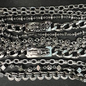 Op de afbeelding: Een assortiment zilverkleurige armbanden met diverse kettingstijlen en decoratieve elementen. De armbanden omvatten gourmetkettingen, schakelkettingen en kettingen met kruis- en bloemmotieven. Sommige armbanden hebben rechthoekige sluitingen met sierlijke patronen.