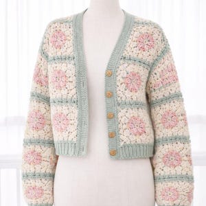Puede incluir: Rebeca color crema con frente abierto y un patrón floral de ganchillo en rosa y crema. Los bordes y puños son de un verde suave, y la rebeca tiene botones de madera. El estilo corto se muestra en un maniquí.