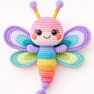 Può includere: Un giocattolo a forma di farfalla all'uncinetto dai colori vivaci, con un viso sorridente e grandi ali multicolori. Il corpo della farfalla è una sfumatura di colori arcobaleno e ha piccole antenne con punte viola. Le ali presentano un design a strati.
