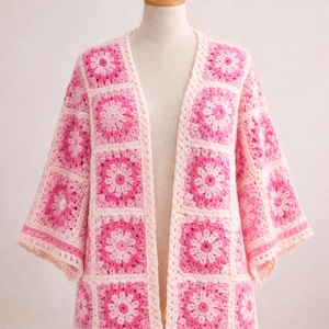 May include: A handmade, open-front cardigan in a granny square design. The crochet jacket features a floral pattern in shades of pink and cream. The sleeves are elbow-length, and the garment is displayed on a mannequin.