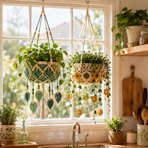 May include: Two hanging macrame plant holders with green plants. One is green and cream with leaf and bead accents. The other is cream with yellow flower accents. Both are suspended in front of a window.