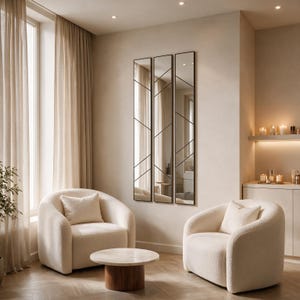 May include: A set of three tall, rectangular wall mirrors with thin black frames and geometric grid patterns, mounted vertically on a light-colored wall above two cream-colored, rounded armchairs and a small round wooden coffee table.
