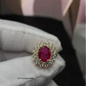 May include: A gold ring featuring a large, oval-cut ruby gemstone surrounded by a halo of smaller, clear gemstones. The ring is held by a gloved hand, showcasing the jewelry's design and craftsmanship. The ring is a statement piece.