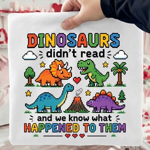 May include: A white sweatshirt featuring a colourful pixel art graphic of four dinosaurs, a volcano, trees, and clouds. The text reads: DINOSAURS didn't read and we know what HAPPENED TO THEM. The design includes small yellow stars and red hearts.