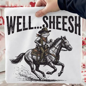 May include: White sweatshirt featuring a cartoon frog in a cowboy hat and vest riding a horse. The text "WELL...SHEESH" is printed above the frog and horse illustration. The frog is wearing glasses and a bandana.