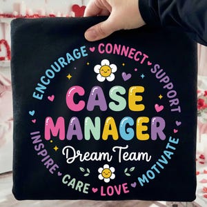 以下が含まれることがあります： カラフルな文字で「CASE MANAGER Dream Team」と書かれた黒い正方形のクッション。デザインにはハート、星、花が含まれており、「Encourage」、「Connect」、「Support」などの言葉がメインテキストを囲んでいます。