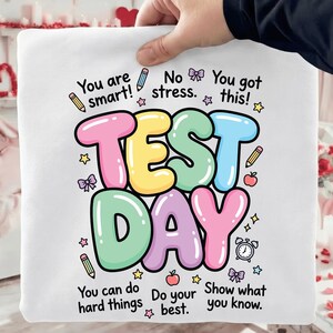 Può includere: Cuscino quadrato bianco con le parole "TEST DAY" in grandi lettere colorate. Il cuscino presenta frasi incoraggianti e illustrazioni di matite, stelle, fiocchi e una sveglia.