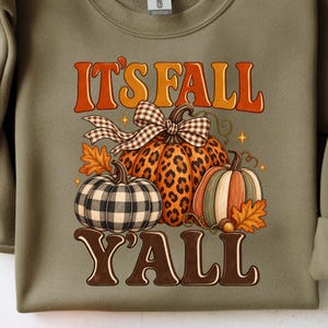Peut inclure: Un sweat-shirt à col rond vert olive avec une impression graphique portant le texte IT'S FALL Y'ALL. Le motif comprend trois citrouilles : une à carreaux noirs et blancs, une à motif léopard et une avec des rayures verticales.