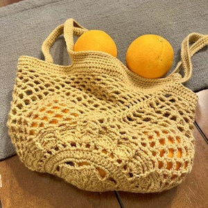 May include: A crocheted, open-weave tote bag in a golden-yellow hue, filled with bright orange fruit. The bag has two handles and a decorative, patterned bottom. The oranges are round and vibrant, contrasting with the bag's texture.